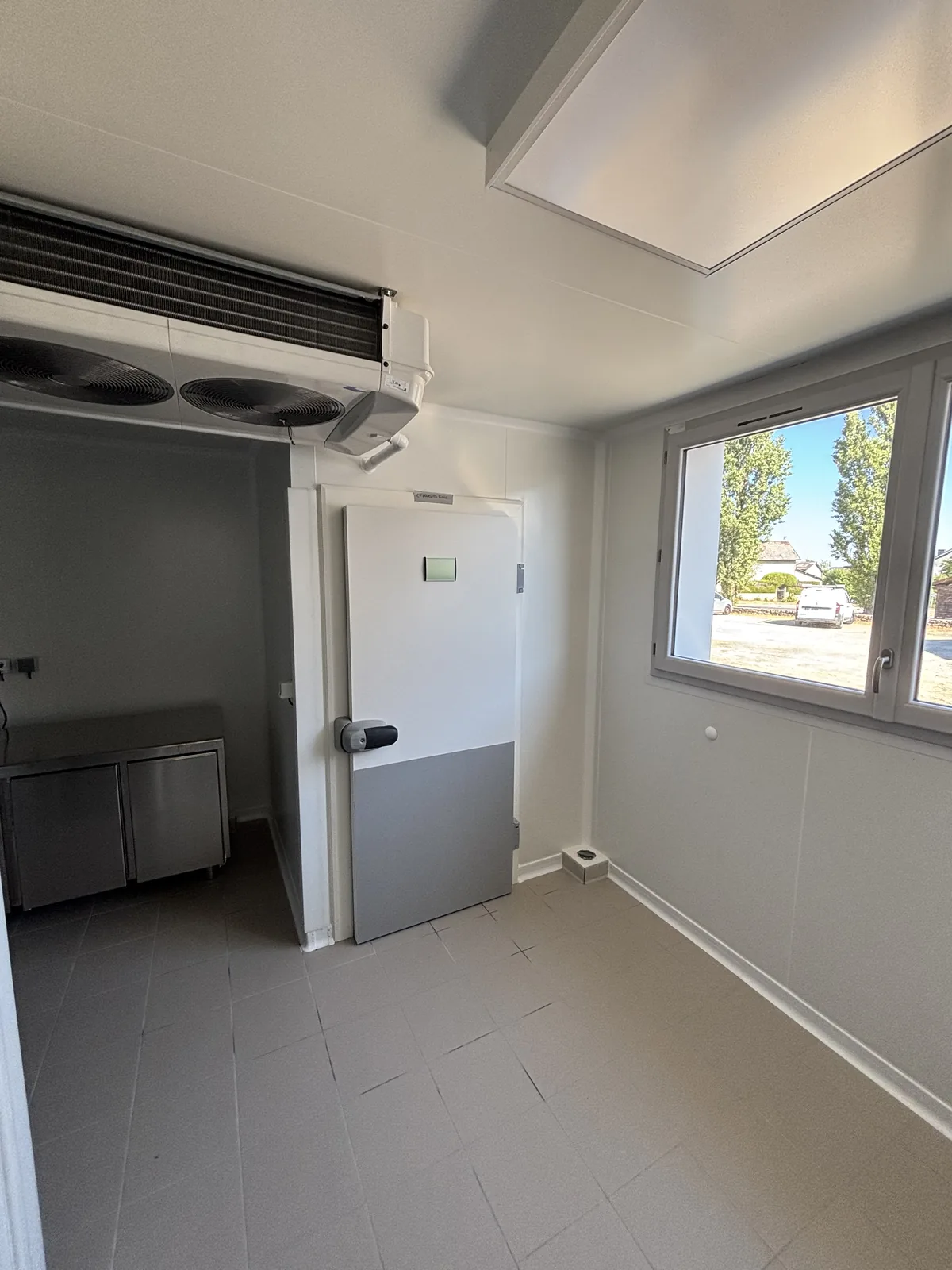 Local réfrigéré d’une cuisine centrale avec évaporateur double ventilateur au plafond et porte pivotante isotherme blanche à plinthe grise