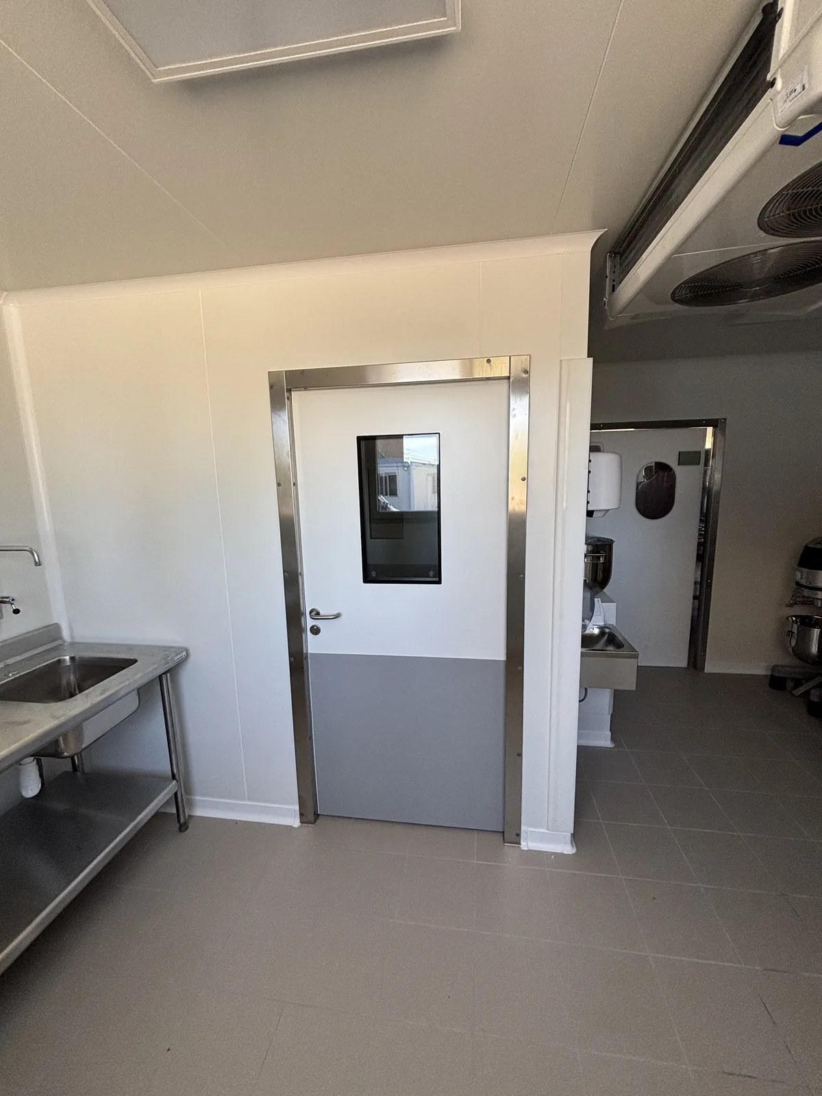 Porte pivotante semi-isolante à oculus et huisserie inox dans une cuisine centrale, évaporateur au plafond et plonge inox à gauche