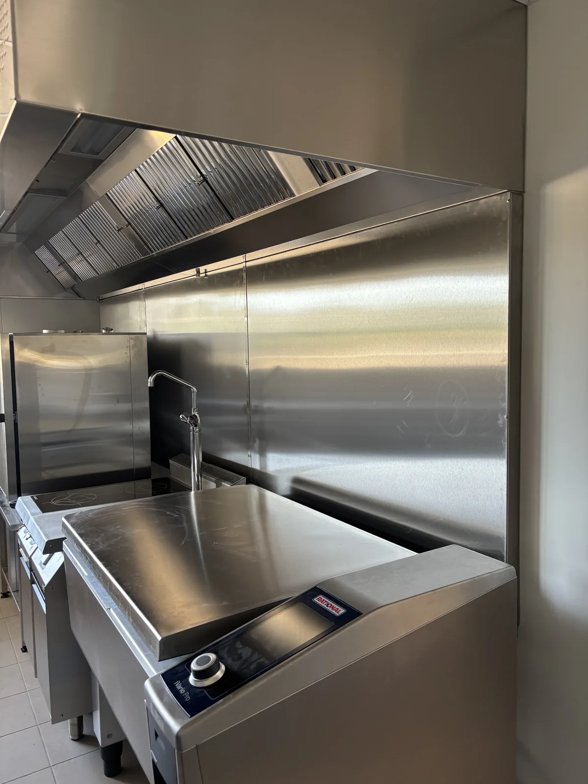 Écran thermique inox posé au-dessus des éléments de cuisson d’une cuisine centrale, sous une hotte à filtres, avec sauteuse et plaque de cuisson